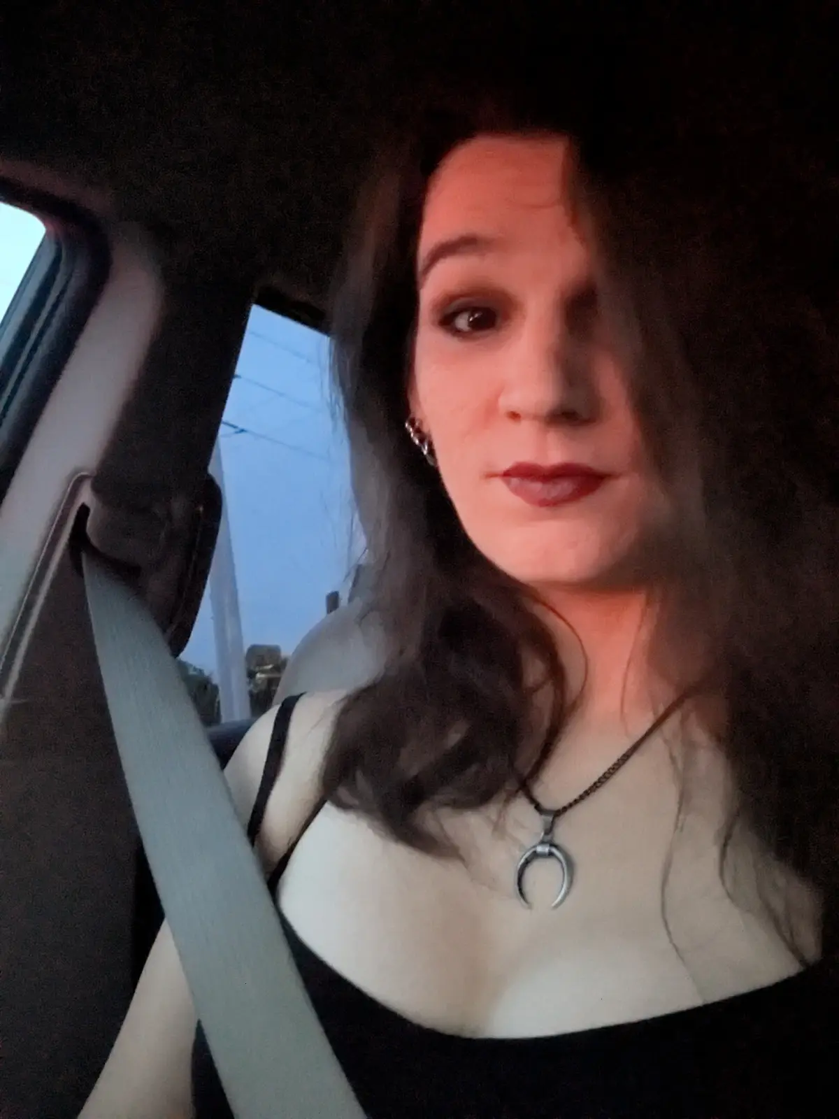 A woman with dark hair and red lipstick sits in a car, wearing a black top and a silver crescent moon pendant necklace, with…