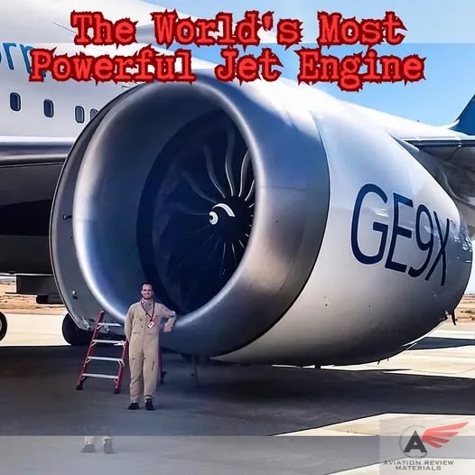 A man in a beige flight suit stands next to a massive jet engine labeled 'GE9X', with a red ladder beside him. The engine is…