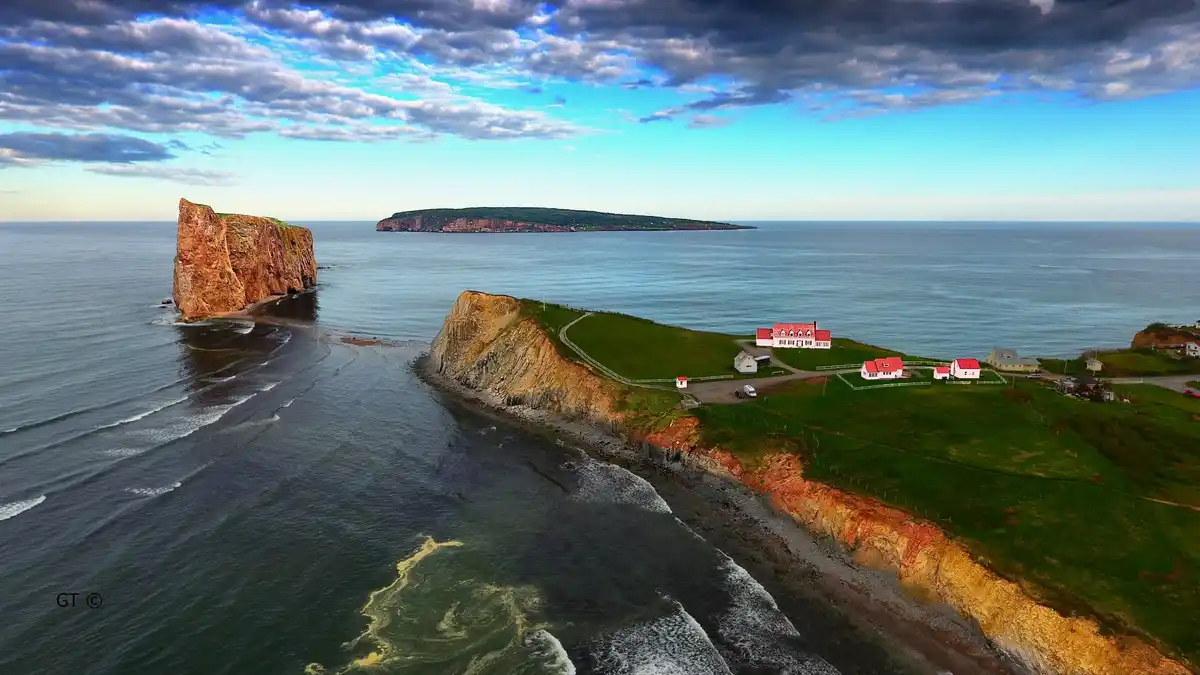 An aerial photograph shows a scenic coastal landscape with dramatic cliffs, ocean waves, and a few white houses with red…