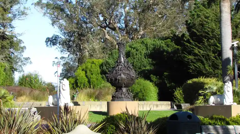 A photograph of a park with a large dark bronze sculpture resembling a vase or urn, flanked by two white stone lion statues…