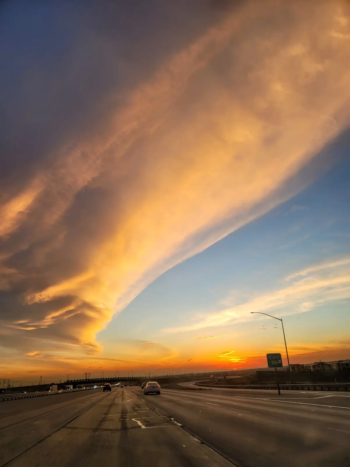 A dramatic sunset on a highway with a massive, fiery-orange cloud formation dominating the sky, casting a warm glow over the…