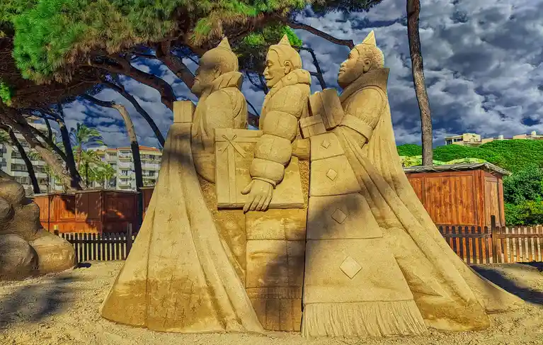 A large, detailed sand sculpture depicts three figures resembling the Three Kings, each holding a gift box, standing under a…