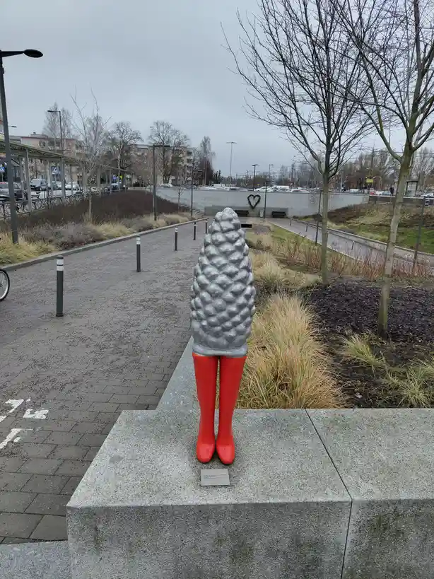 A sculpture of a person viewed from behind, featuring a silver, pinecone-shaped torso and bright red boots, standing on a…