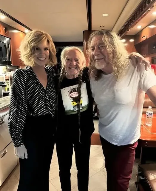 Three people pose together inside a luxurious RV kitchen. The woman on the left wears a black and white polka dot blouse and…