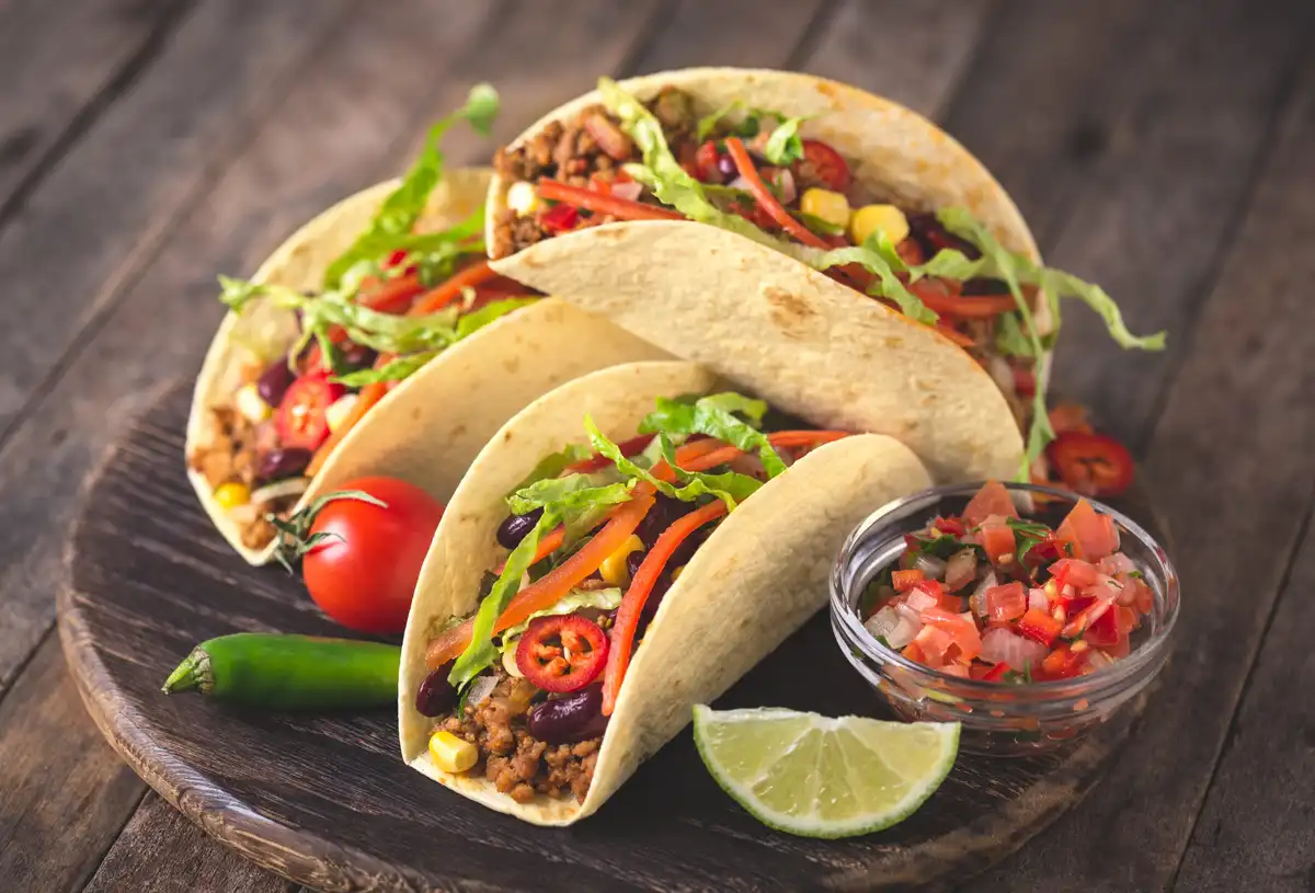 Three soft-shell tacos filled with ground meat, lettuce, corn, beans, and peppers sit on a dark wooden plate, accompanied by…