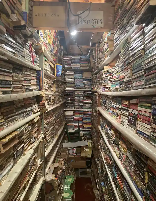 A narrow aisle in a cluttered used bookstore is flanked by towering shelves packed with books, with a handwritten sign above…