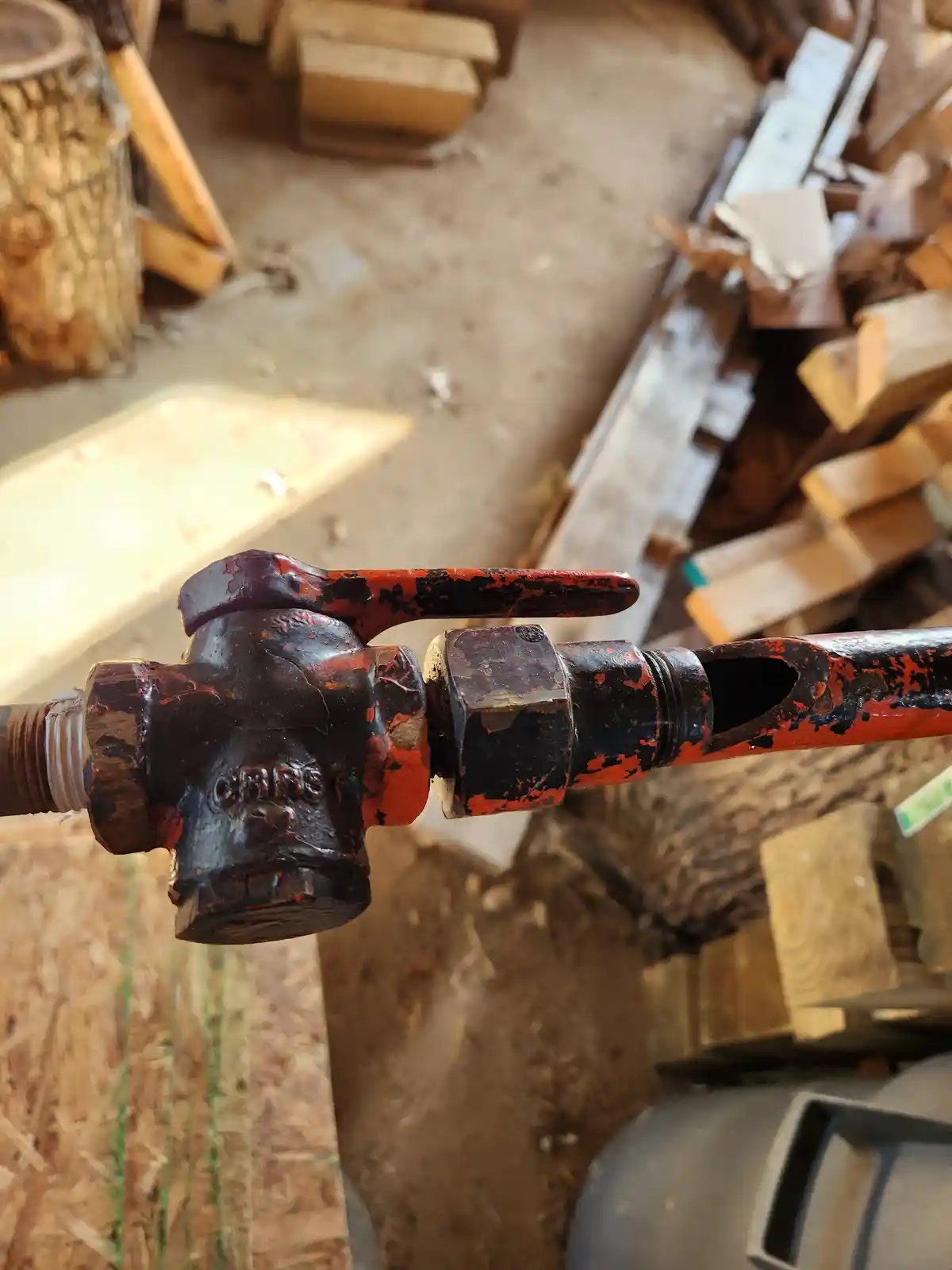 A close-up of a weathered, red-and-black painted valve handle in a cluttered workshop, with wood piles and tools in the…