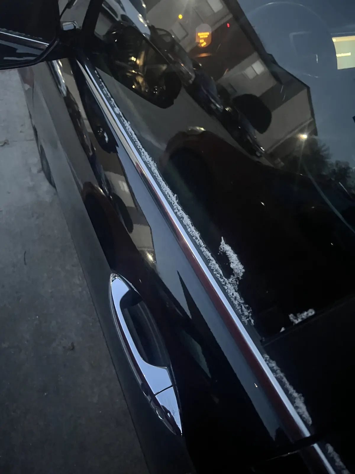 A close-up, angled shot of a black car parked at night, with frost or snow visibly accumulating along the window line and…