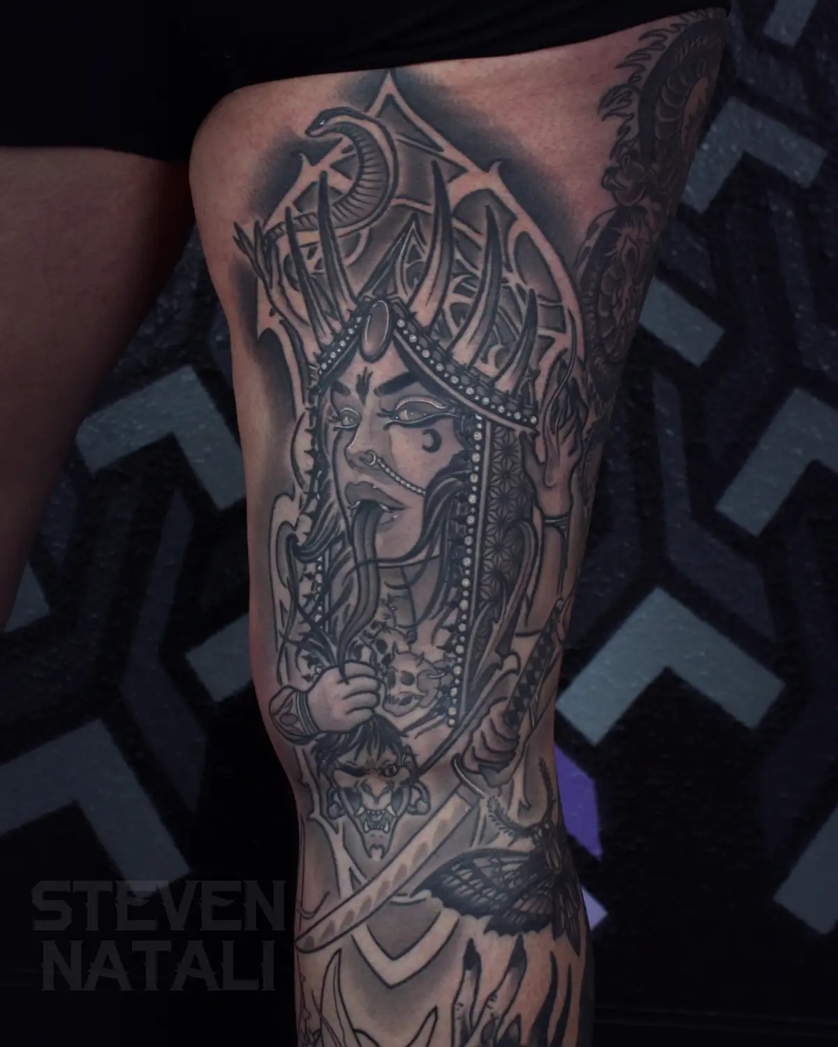 A close-up photo shows a detailed black and grey tattoo on a person's thigh, depicting a stylized, horned deity with…