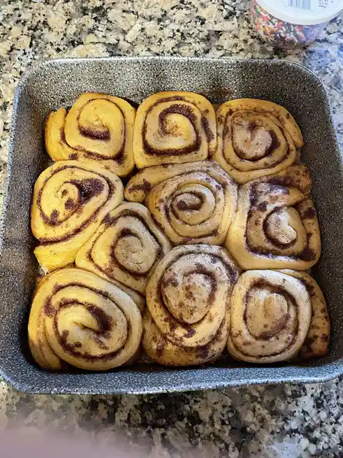 A close-up photo shows freshly baked cinnamon rolls arranged in a square metal baking pan on a granite countertop. The rolls…
