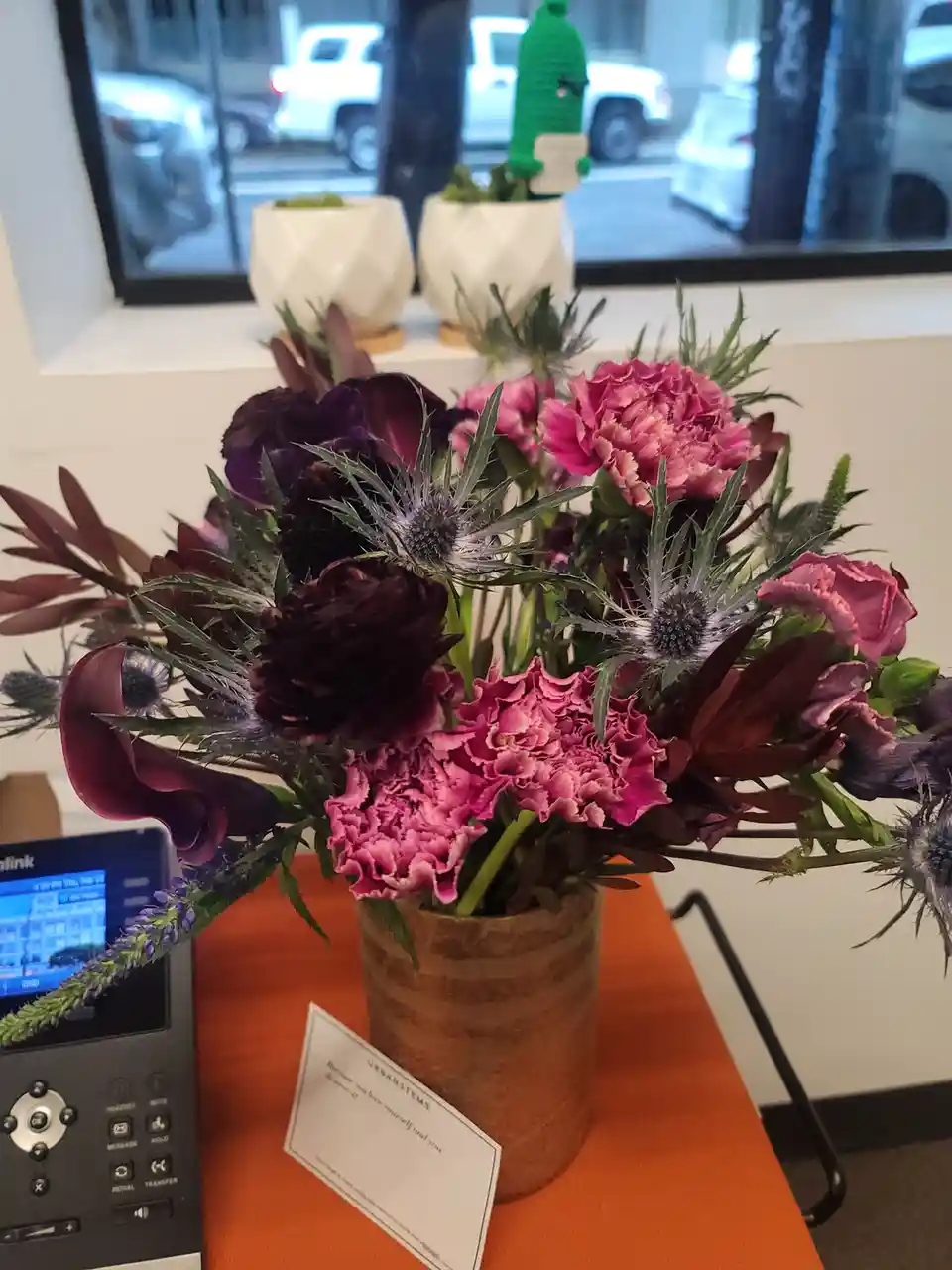 A vibrant floral arrangement with pink carnations, dark purple flowers, and spiky thistles sits in a brown vase on an orange…