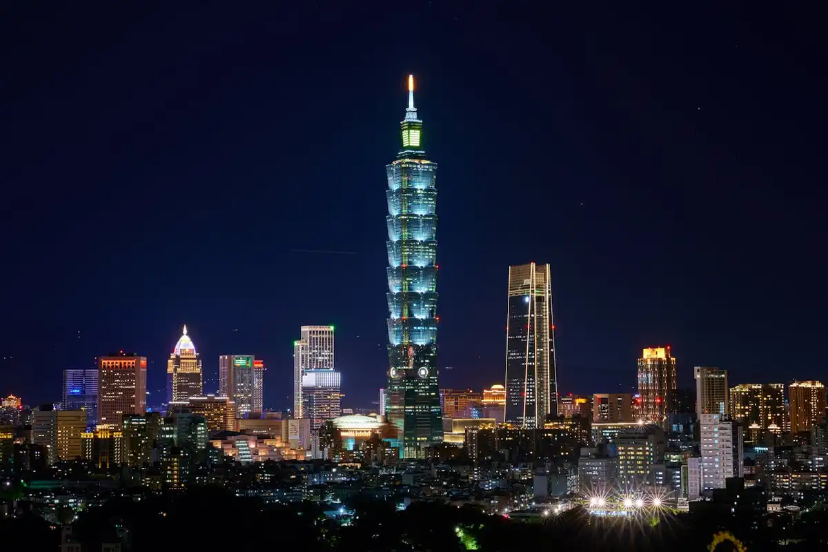 A nighttime cityscape featuring the illuminated Taipei 101 skyscraper as the central focus, surrounded by other lit…