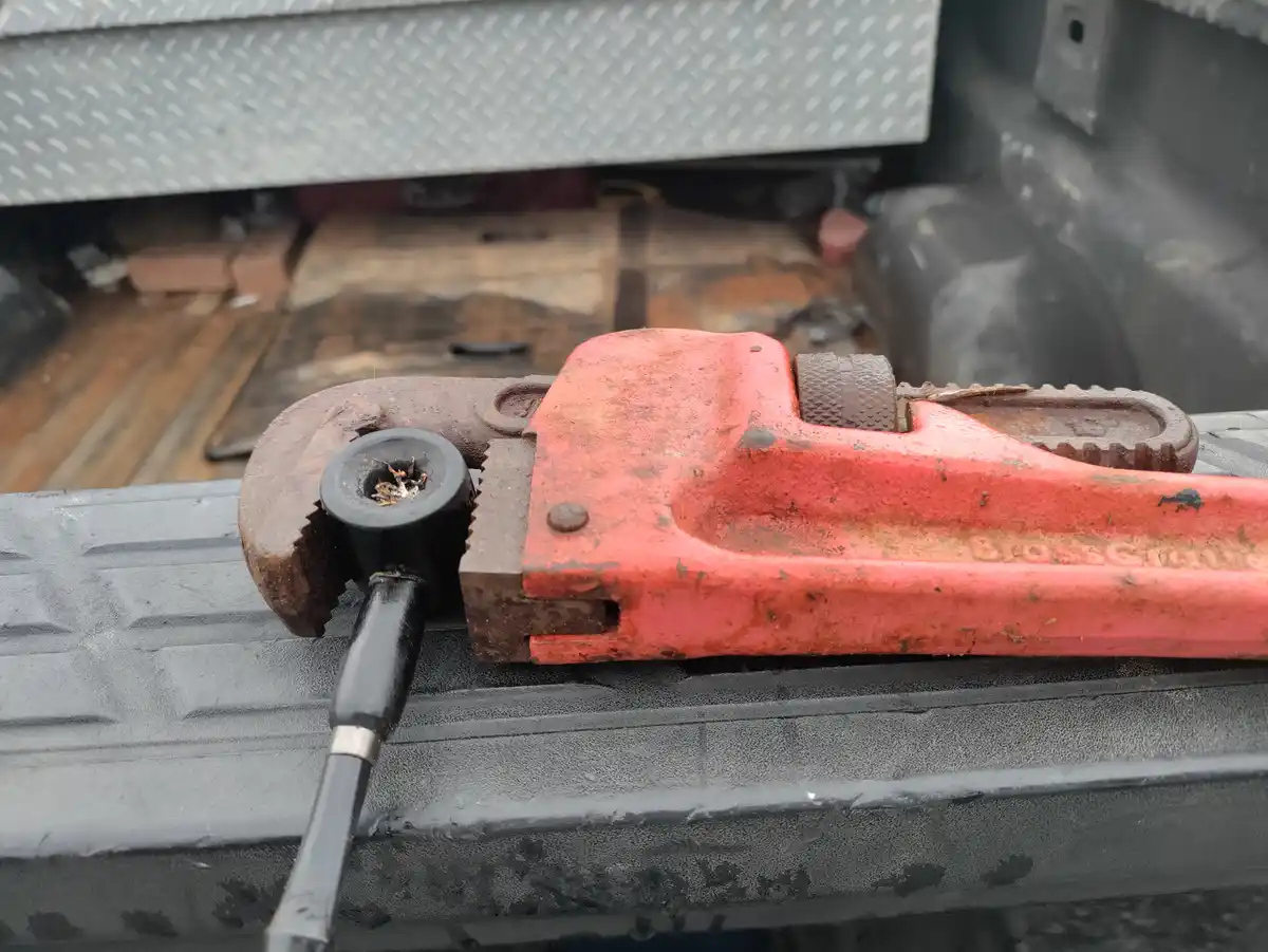 A red, rusty pipe wrench sits on the edge of a truck bed, its jaws holding a black pipe smoking with a small fire inside…