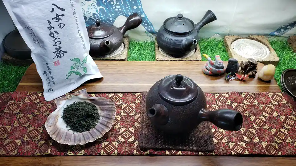A still life of traditional Japanese tea culture featuring three dark ceramic teapots on wooden trays, a bag of Japanese…