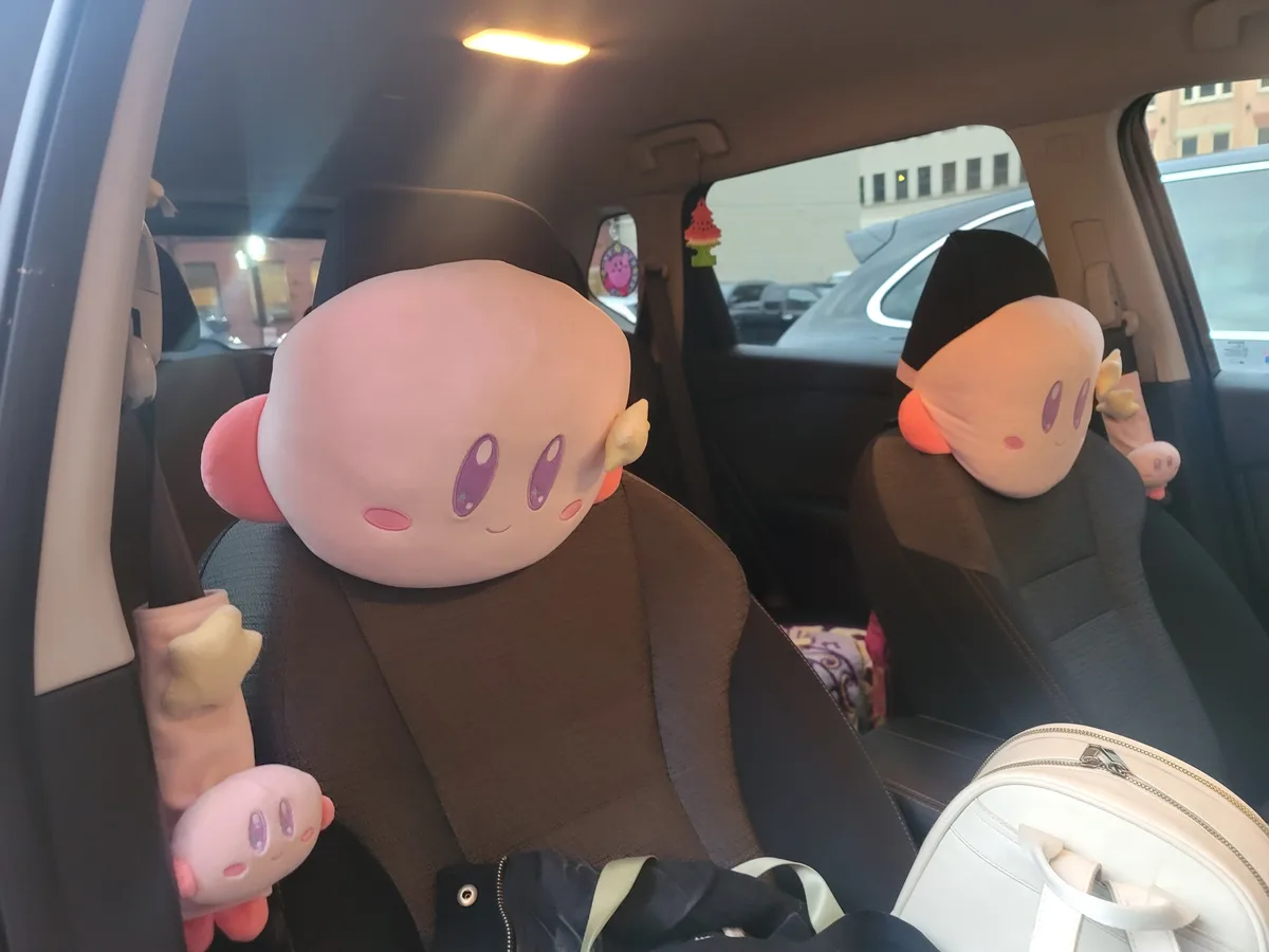 Inside a car, two large Kirby plushies are strapped to the headrests of front seats, with smaller Kirby plushies hanging…