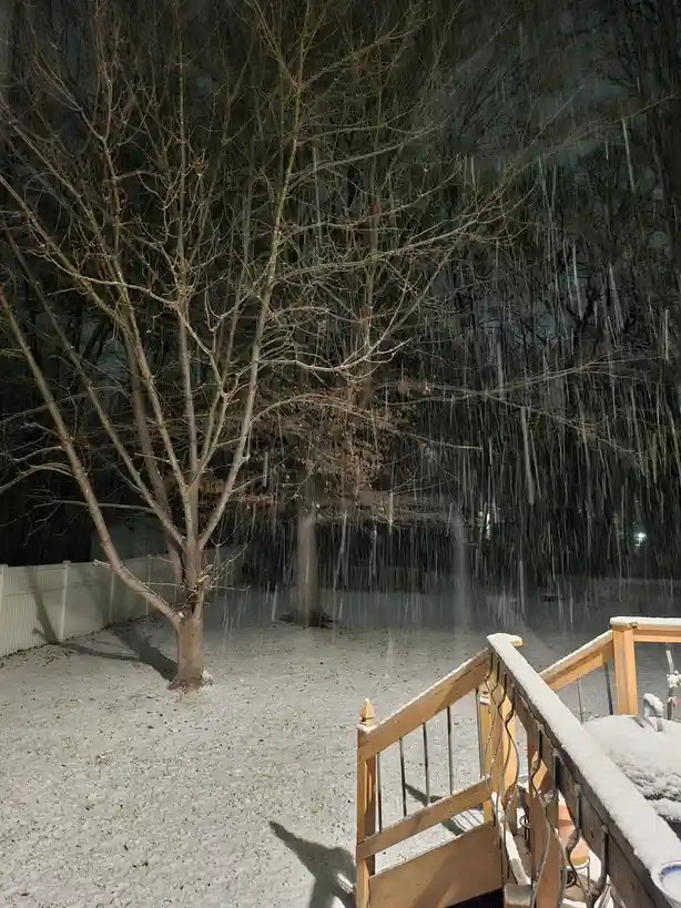 A nighttime backyard scene shows bare trees covered in long, dangling icicles, with snow on the ground and a wooden deck…