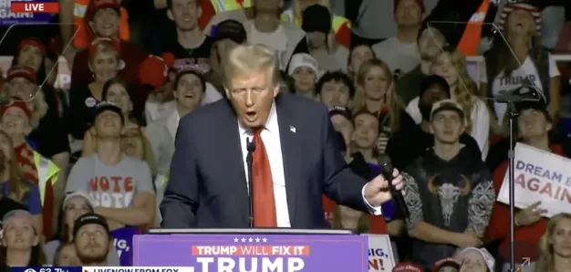 A man in a dark suit and red tie stands at a purple podium with the text 'TRUMP WILL FIX IT' on it, shouting with his mouth…