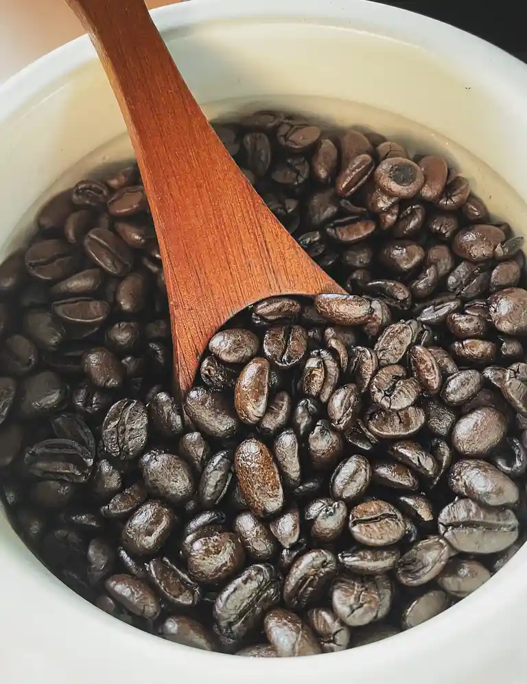 A close-up photo shows a white ceramic bowl filled with dark roasted coffee beans, with a wooden spoon resting inside…