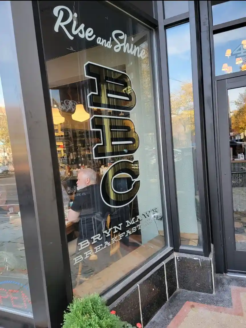 A storefront window displays the text 'Rise and Shine BBC Bryn Mawr Breakfast Club' in stylized lettering. A man with a…