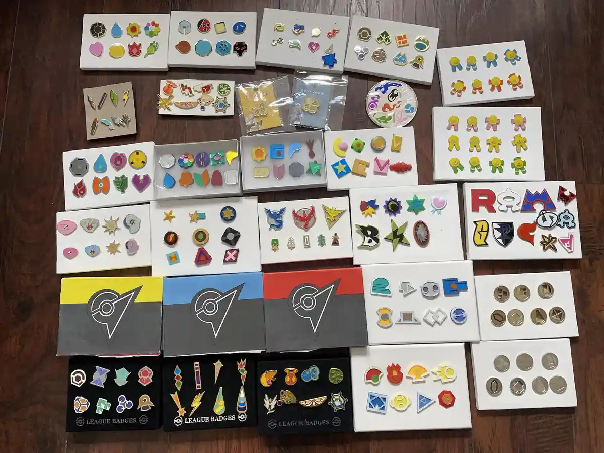 An overhead shot displays dozens of Pokémon League Badges arranged on white boards and in boxes on a wooden floor. The…