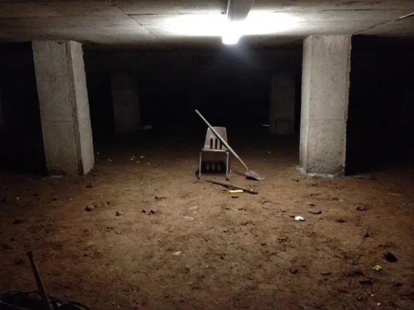 A dimly lit, dusty basement with concrete pillars and a single overhead light illuminating a plastic chair, a broom leaning…