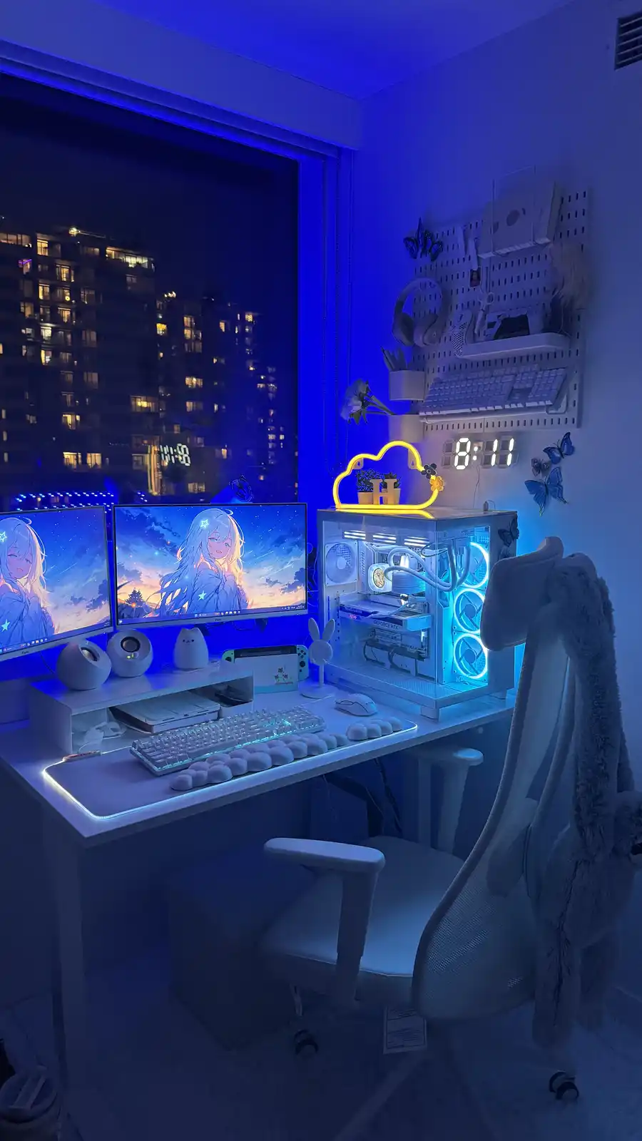 A meticulously arranged, blue-lit gaming or streaming setup at night, featuring two monitors displaying anime girl…