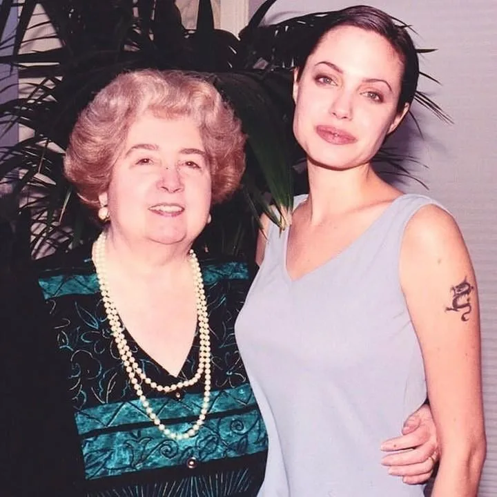 An older woman with curly blonde hair and pearl necklaces stands beside a younger woman with dark hair and a grey dress, who…