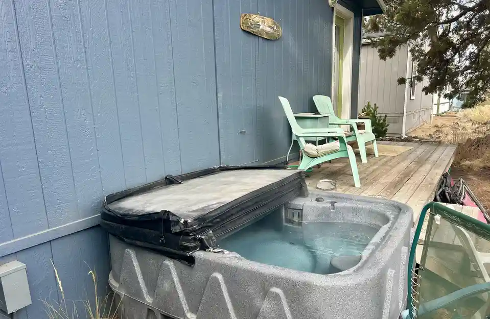 An outdoor hot tub with its black cover partially lifted, revealing warm water inside, situated on a wooden deck next to a…