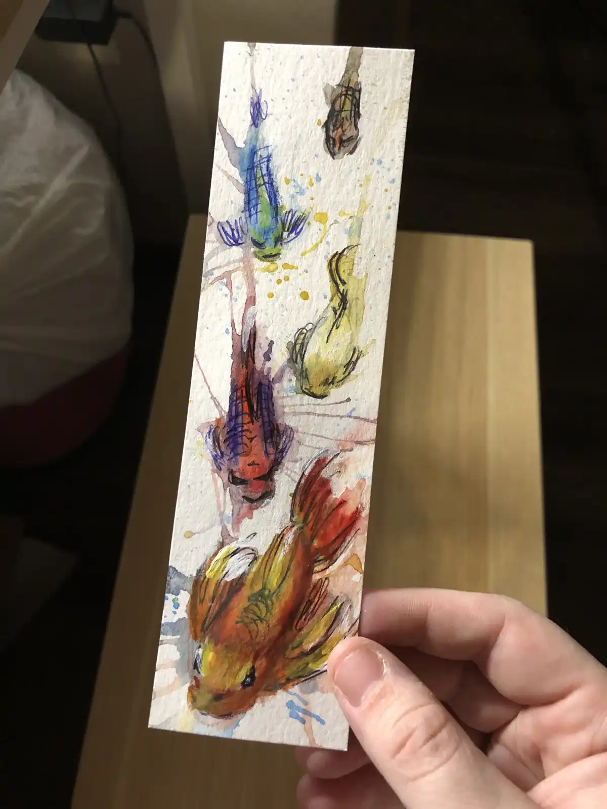 A hand holds up a small, vertically oriented watercolor painting on textured paper depicting several stylized, colorful fish…