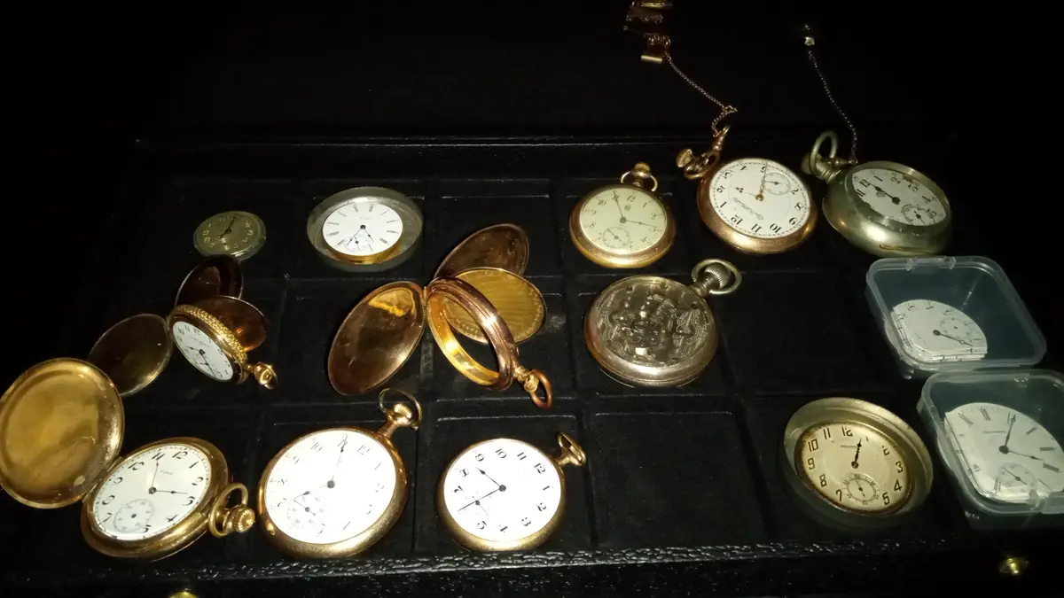 A collection of vintage pocket watches in various gold and silver tones are displayed in a black velvet-lined case, some…