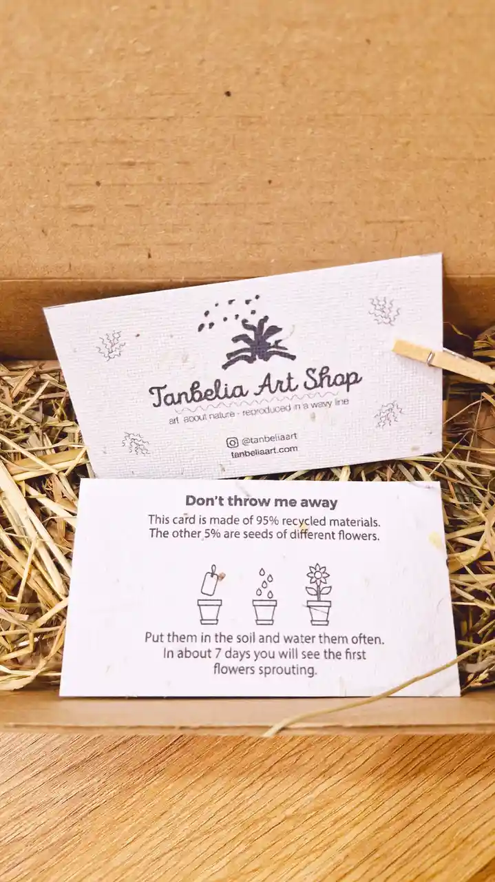 Two white cards rest on straw inside a cardboard box. The top card is branded 'Tanbelia Art Shop' with social media info…