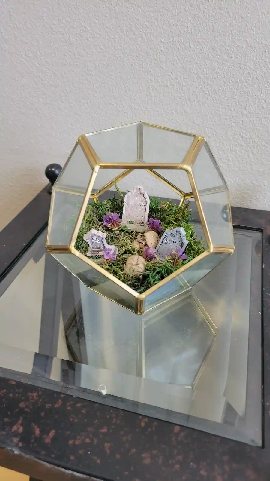 A hexagonal glass terrarium with gold trim sits on a mirrored table, containing green moss, purple flowers, miniature…
