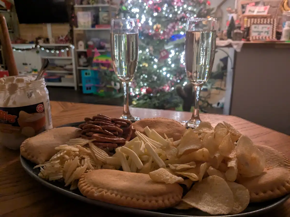 A wooden table holds a plate of assorted snacks including potato chips, pretzels, crackers, and empanadas, with two glasses…