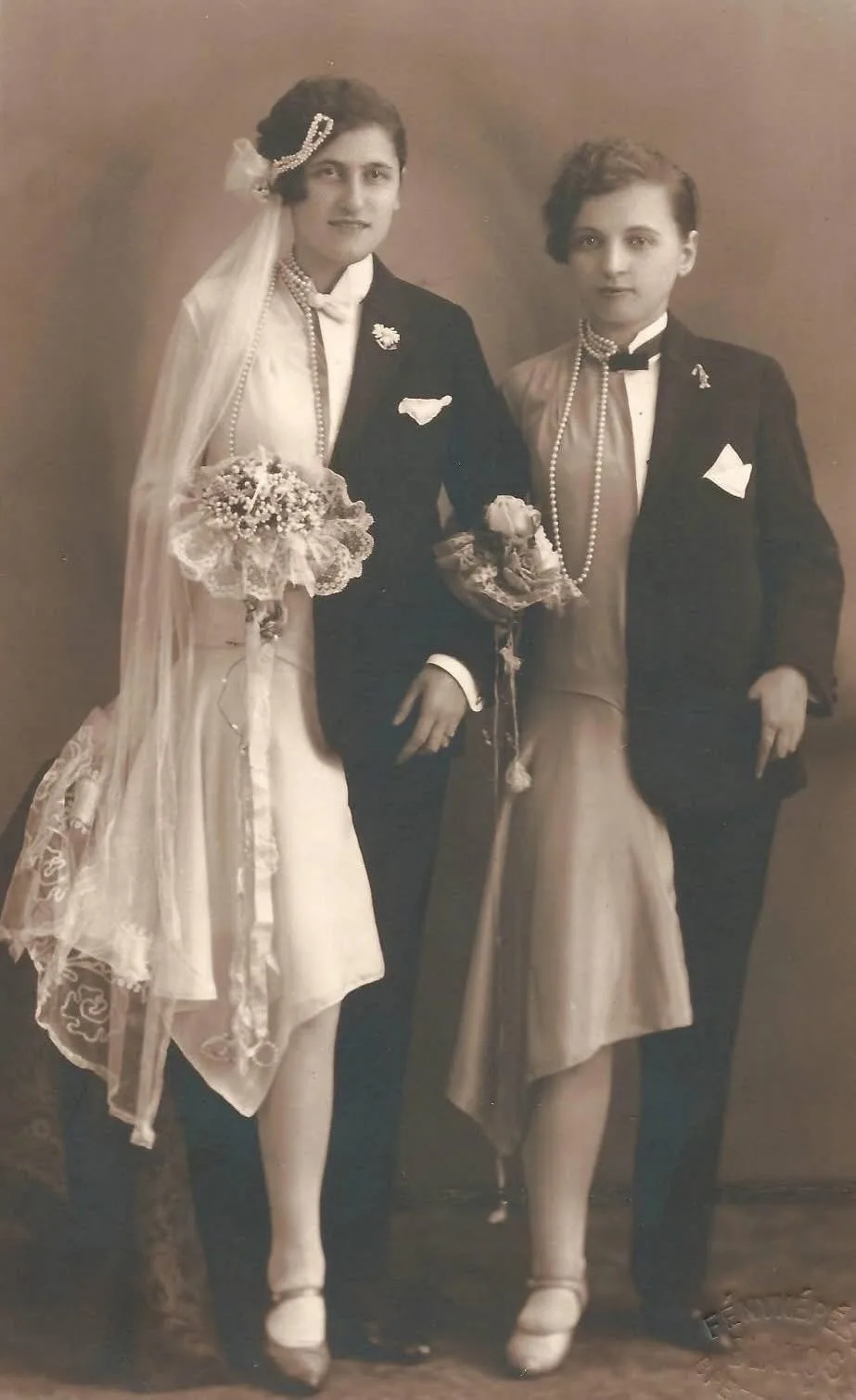 A vintage sepia-toned wedding photo shows two women dressed in formal attire, one wearing a veil and holding a bouquet, both…