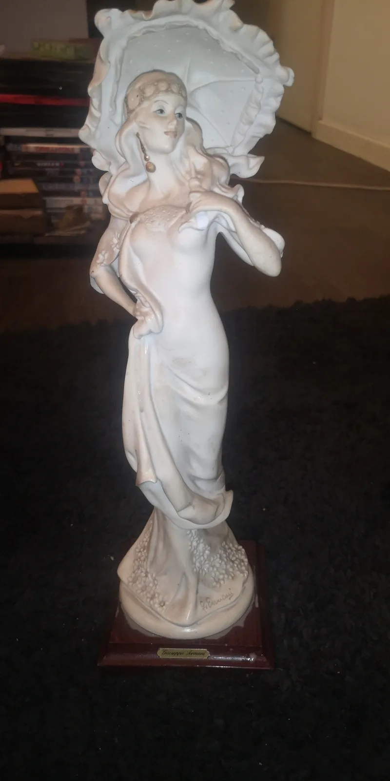 A white ceramic statue of a woman in a flowing dress holding a large parasol, standing on a wooden base with a plaque that…