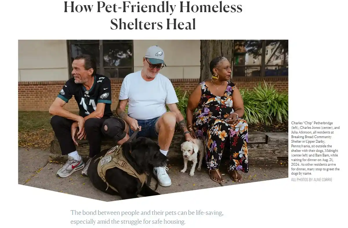 Three people sit on a bench outside a shelter with two dogs, one large black service dog wearing a vest and a small white…