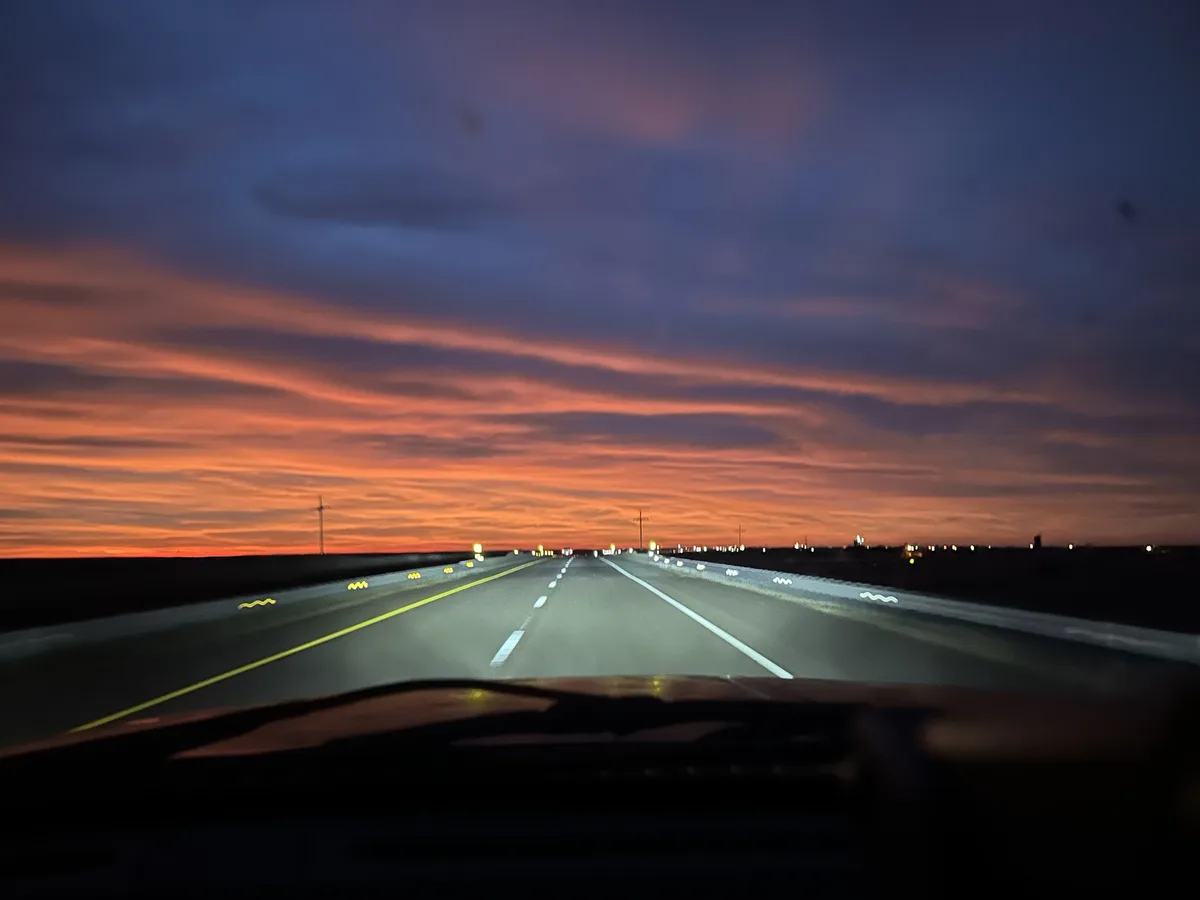 A photo taken from inside a car looking forward at a highway at dusk, with the sky painted in vibrant orange and purple…