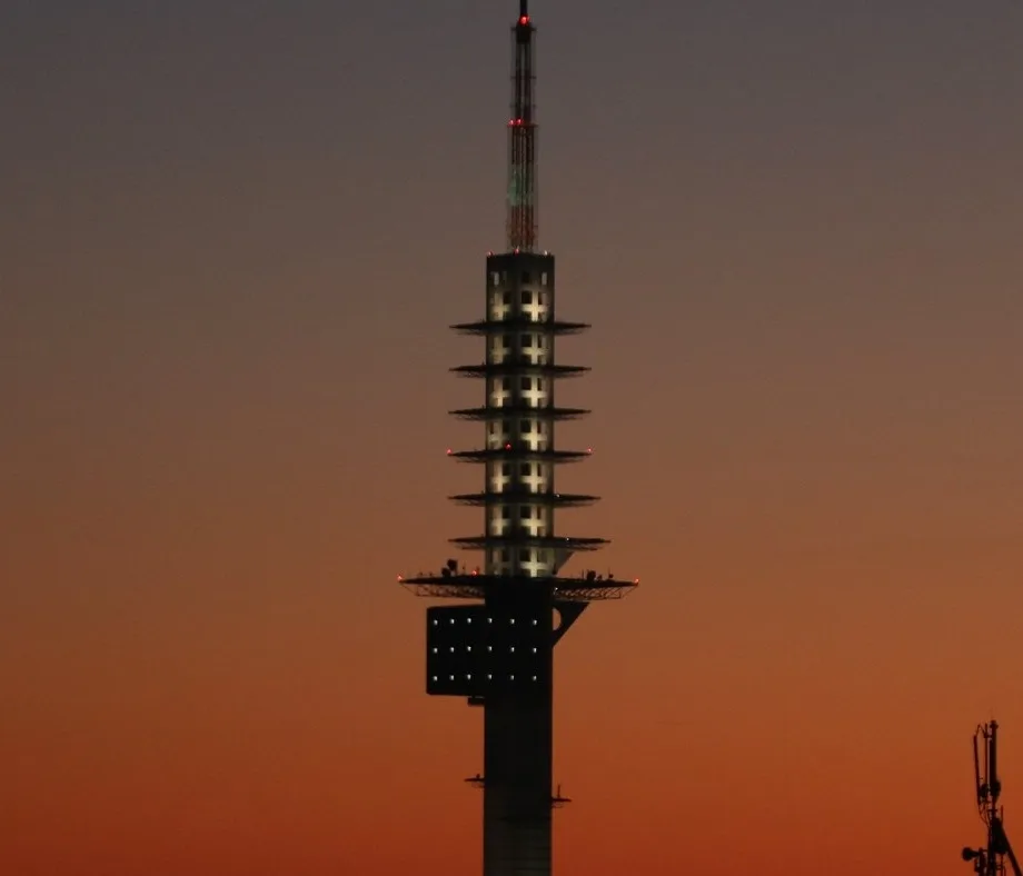A tall, modern telecommunications tower stands against a twilight sky with orange and purple hues. The tower has multiple…