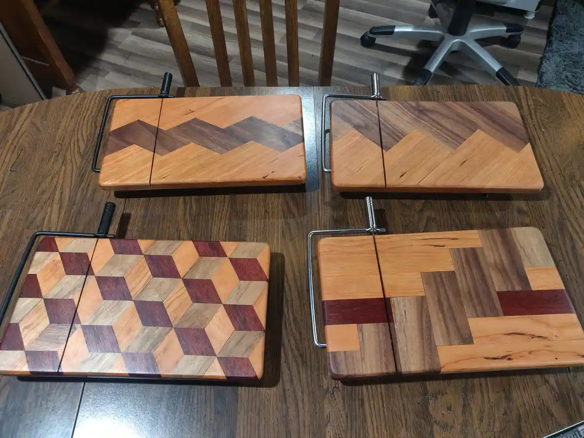 Four wooden cutting boards with intricate geometric inlays are displayed on a dark wood table. Each board has a unique…