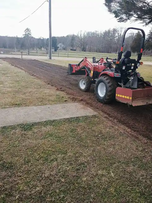 A red and black compact tractor with a front loader and rear blade is parked on a dirt path next to a concrete sidewalk…
