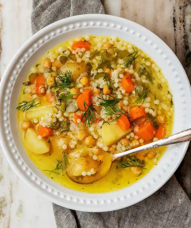 A top-down photo of a white bowl filled with a hearty vegetable soup containing chickpeas, pearl couscous, diced carrots…