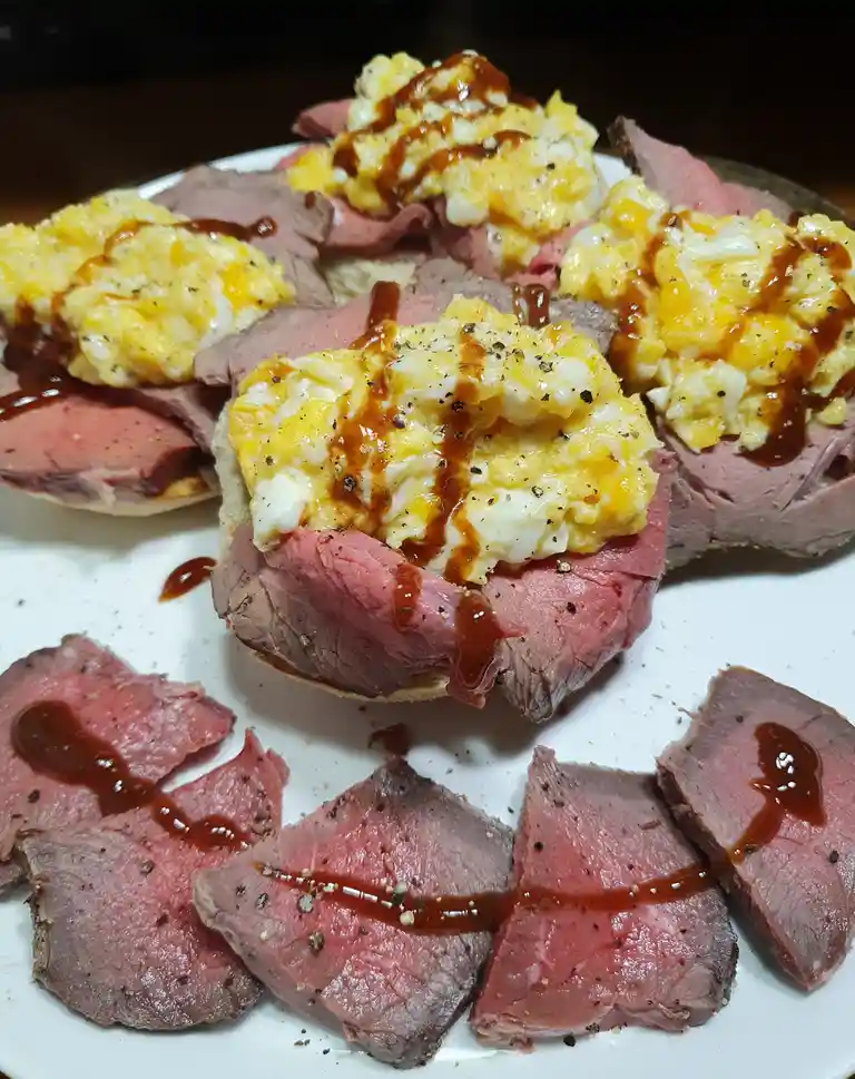 A plate of sliced rare beef, some arranged on bread with scrambled eggs and drizzled sauce, others scattered around. The…