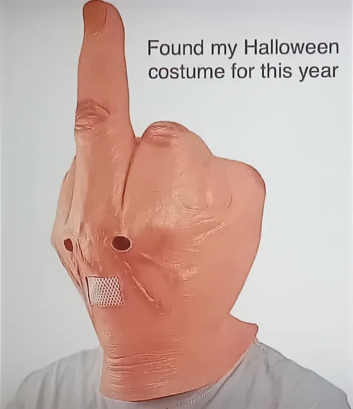 A person wears a large, flesh-toned rubber mask shaped like a hand making a rude gesture, with holes for eyes and a mesh…