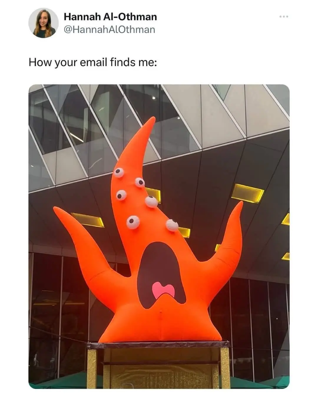 An orange inflatable monster with multiple googly eyes and a wide-open mouth stands on a pedestal in front of a modern glass…