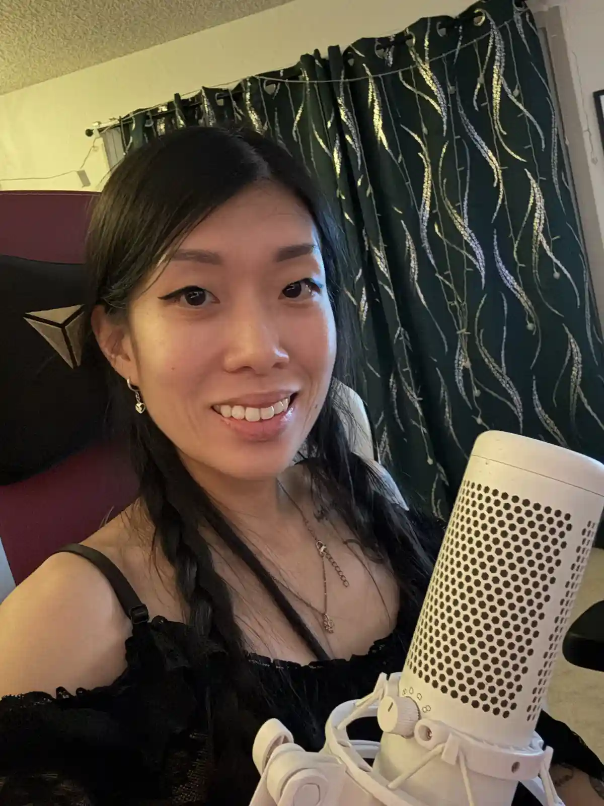 A woman with long dark hair and a black top smiles at the camera while sitting in front of a white condenser microphone…