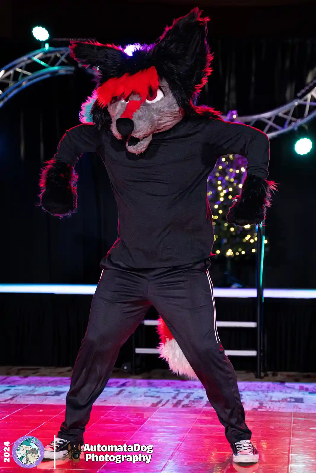A person in a black and red fox fursuit strikes a dynamic dance pose on a stage lit with pink and purple lights, with a…