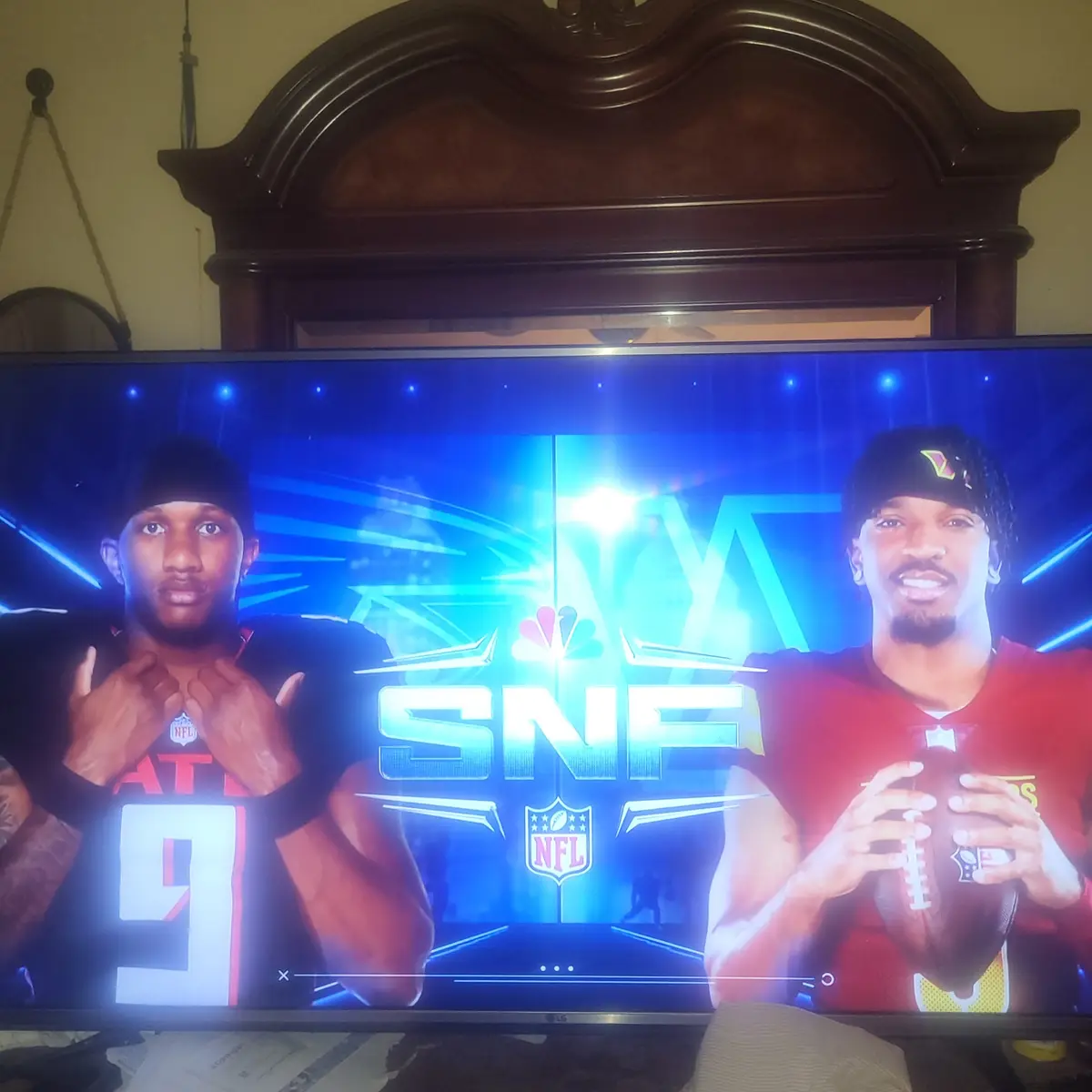A television screen displays the 'SNF' (Sunday Night Football) intro graphic with two NFL players, one in a black jersey…