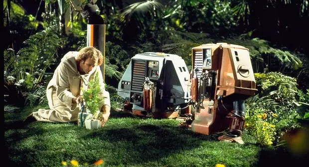 a man in a beige robe kneels in a lush green garden, tending to a small potted plant. Two droids, one gray and one brown…