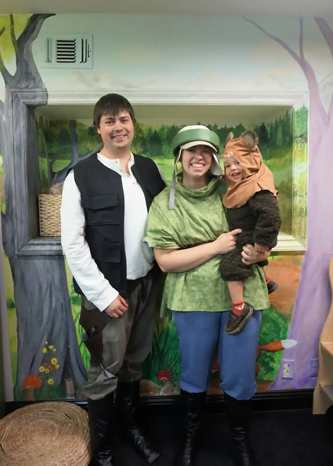 A family of three poses in front of a painted forest backdrop, dressed as characters from Star Wars: the man wears a black…