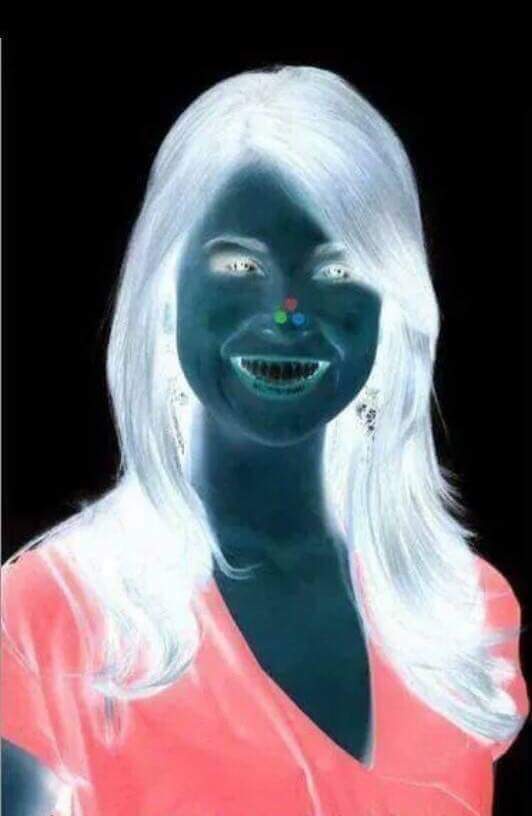 A woman with glowing white hair and a dark blue skin tone smiles widely, revealing teeth, while wearing a pink top against a…